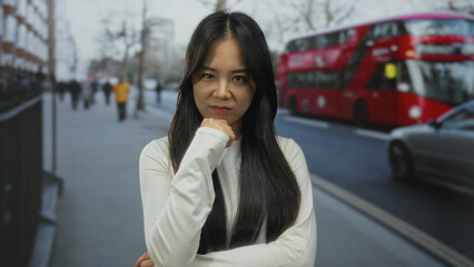 Fototapeta premium Woman standing on a street in london with red bus and cars passing by, wearing white shirt, expressing confidence and contemplation in an urban outdoor setting.