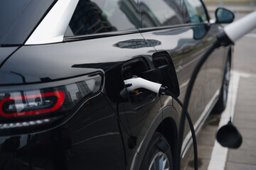 A black electric car is plugged into a charging station located in a busy urban setting. The charging process is highlighted by the sleek design of the vehicle and station.