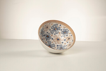 Close-up of a decorative ceramic bowl resting at an angle on a white surface. The interior features a hand-painted floral pattern in blue and tan tones, framed by a woven-style rim.
