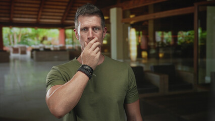 Fototapeta premium Man with hand to mouth gesture in hotel building lobby, wearing green shirt and bracelets; worry introspection.