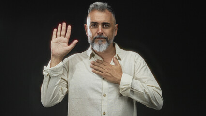 Man with hand on chest and raised palm in black studio, bearded middle age male with eyes closed in solemn pledge pose; sincerity calm.