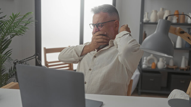 Man yawning with hand over mouth and stretching at laptop on desk amid lamp and shelving in building home office; tiredness remote work.