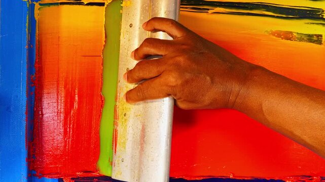 A close up view of hands carefully blending yellow, orange, and red inks on a screen printing frame, creating a vibrant gradient effect. highlighting the meticulous artistry of manual printmaking.