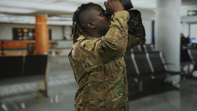 Man in camouflage military uniform looking through binoculars at airport terminal indoors; vigilance duty. - Powered by Adobe