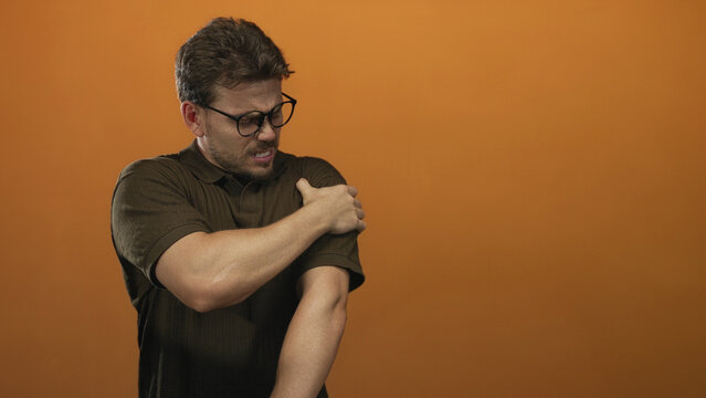Young hispanic man clutching shoulder with pained face in studio showing shoulder pain with orange backdrop; pain recovery.