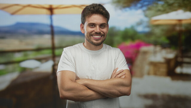 Young man smiling with arms crossed and bare forearms on restaurant terrace building outdoors; confidence.