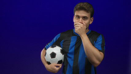 Young hispanic man holding a soccer ball under his arm, hand covering mouth, wearing blue and black striped jersey in a blue studio set; amused hush.