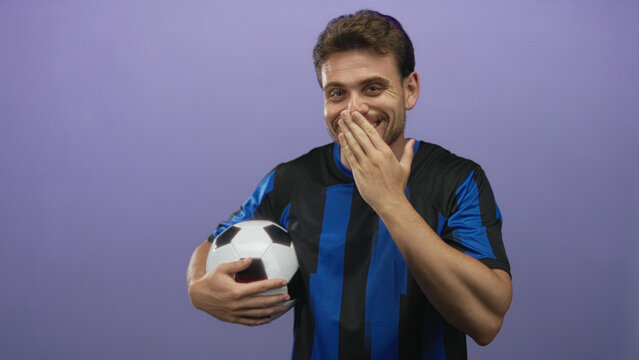 Young hispanic man holding a soccer ball with hand over mouth gesture in studio with purple background; playful.