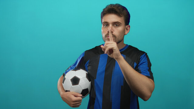Young hispanic man holding soccer ball with index finger to lips gesture in teal studio; silence focus.