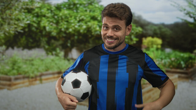 Young man in blue striped jersey smiling while holding a soccer ball in a forest park outdoors with hand on hip; confidence team spirit.
