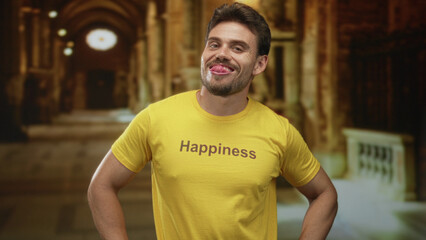 Fototapeta premium Man smiling with hands on hips wearing yellow tshirt printed with the word happiness in a church aisle with arches and railing; joy.
