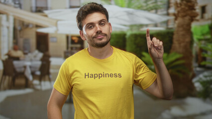 Fototapeta premium Man smiling and giving thumbs up on restaurant terrace wearing a yellow tshirt with the word happiness visible; optimism hope.