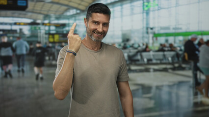 Man points index finger up in airport terminal showing hand and forearm while smiling to camera; optimism travel ease.