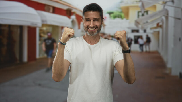 Hispanic man raising both fists and smiling while standing center frame on a sunlit street lined with shops and awnings; joy victory celebration.