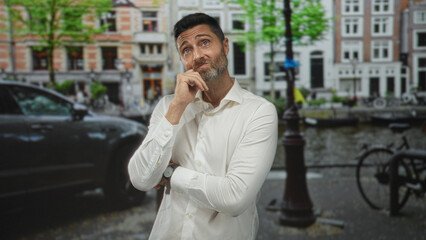 Man with hand on chin and crossed arms standing on canal street in amsterdam next to bicycle and lamppost, thoughtful squinting expression; pensive contemplation.