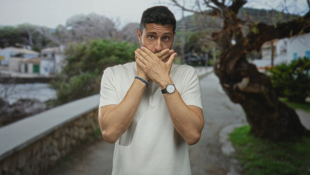 Man covers mouth with hands on a seaside street promenade beside a stone wall and trees; surprise reflection. - Powered by Adobe
