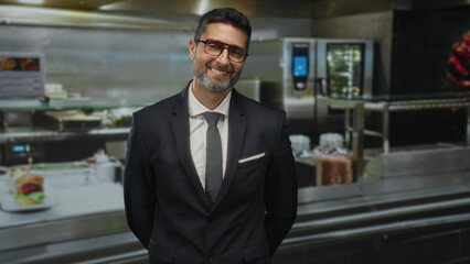 Man with visible face and beard smiling, wearing suit and tie, hands behind back in commercial kitchen; business confidence leadership.