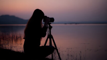 A photographer's silhouette kneels by a calm lake at twilight, focused on capturing wildlife or the serene landscape using a camera mounted on a tripod.