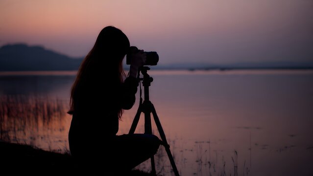 A photographer's silhouette kneels by a calm lake at twilight, focused on capturing wildlife or the serene landscape using a camera mounted on a tripod.