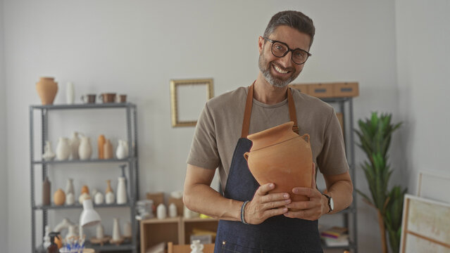 Man smiling and holding a terracotta jar with both hands in studio surrounded by pottery shelves and tools; calm contentment.
