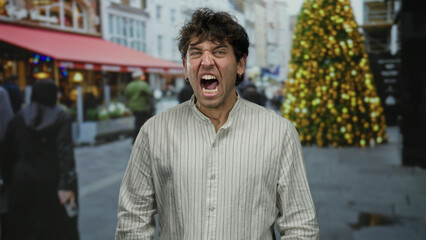 Fototapeta premium Hispanic man expressing emotion outdoors on a bustling street with a festive atmosphere, surrounded by blurred people and a decorated tree in the background.