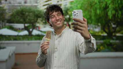 Man enjoying ice cream while smiling during a video call in a vibrant green park setting.