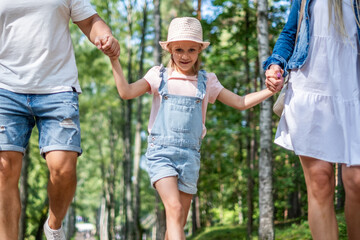 Fototapeta premium Father, mother and daughter walking hand in hand through a lush green forest, enjoying playful moments together, celebrating family bonds and childhood happiness