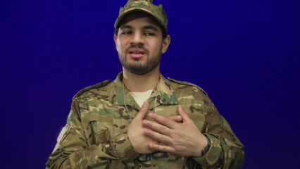 Man in camouflage uniform clutching chest with closed eyes in blue studio wearing metal dogtags;...