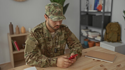 Man soldier in camouflage uniform wearing dogtags seated at wooden table looking at phone in living room; introspection solitude reflection concentration.