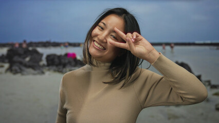 Young woman gesturing peace sign by the seaside on a beach with rocks in the background, showcasing a joyful moment outdoors.
