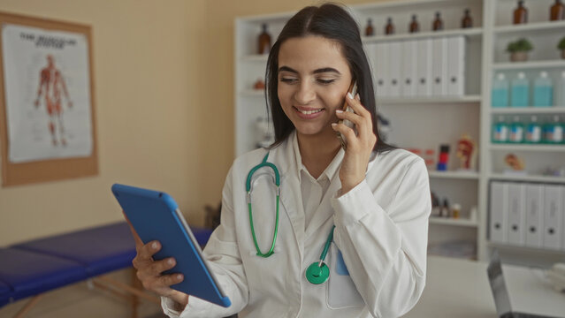 Hispanic woman doctor in uniform with stethoscope talking on phone while using tablet in clinic room showcasing efficient multitasking in healthcare workplace. - Powered by Adobe