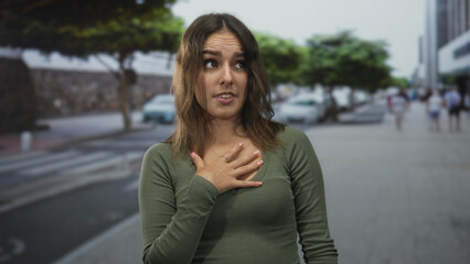 Fototapeta premium Young woman with hand on chest wearing green shirt and necklace on city street pedestrian walkway; regret apology.