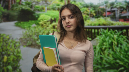 Fototapeta premium Woman holding notebooks and gripping a yellow journal while wearing a backpack on a park path with green foliage and wooden railing; study focus contemplation.
