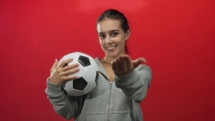 Woman holding a soccer ball and offering her outstretched hand in a red studio setting, smiling; confidence.
