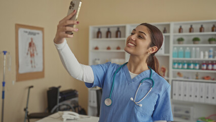 Young woman doctor taking a selfie in a hospital room wearing scrubs and stethoscope, smiling...
