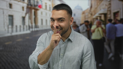 Man with finger to lips for silence in a crowded european city street lined with historic buildings and blurred pedestrians; hush.