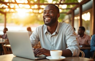 Obraz premium Smiling Black man types on laptop at cafe table with coffee. He works remotely outdoors, happy with his freelance job. Young professional enjoys life.