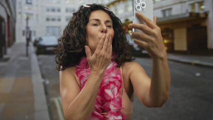Middle age hispanic woman in pink bikini blows kiss to camera while holding smartphone on a busy city street; affection flirtation joy.