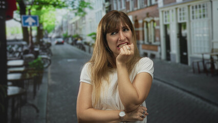 Woman biting nails with hand at mouth on a cobblestone street lined with bicycles and brick buildings; anxiety worry.