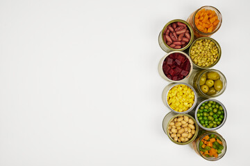 Open cans of beans, mushrooms, carrots, corn, peas, green beans, lentils, asparagus, and olives on a white background with copy space.