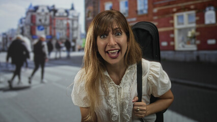 Young woman holding guitar case with hand on chest, looking startled on a busy city street crosswalk near storefront; surprise concern.