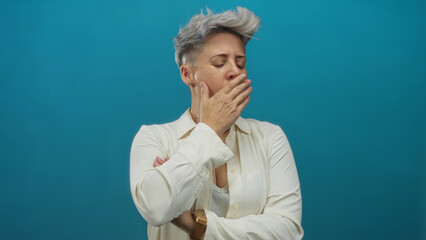 Caucasian woman with short grey hair yawning with hand over mouth in studio; exhaustion and fatigue.