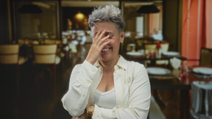 Smiling woman with short hair covers face with hand in building restaurant interior; lighthearted amusement.