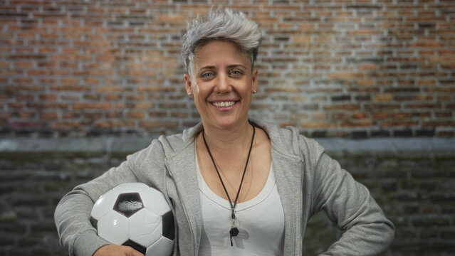 Woman coach holding soccer ball under arm while smiling at brick building with whistle around neck; teamwork spirit confidence.