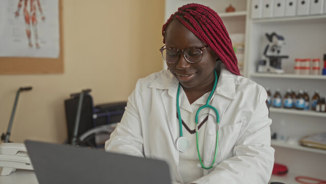 Woman doctor with glasses and stethoscope working on laptop in clinic office surrounded by medical equipment and anatomy chart providing healthcare services indoors for patients.