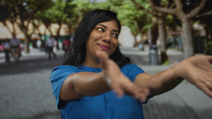 Young african american woman in blue shirt extends arms toward camera on a tree lined city street; joyful welcome.