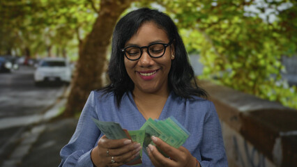 Woman holding and counting green ringgit bills with both hands on city street under trees;...