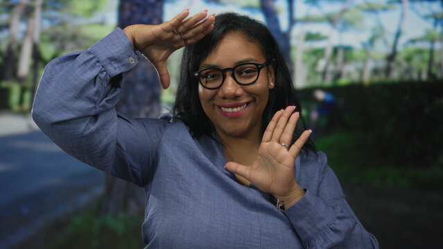 African american woman smiling behind eyeglasses framing face with hands in sunlit green forest; creative joy.