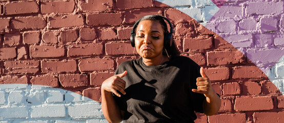 African american woman dancing playfully with headphones against painted wall, banner with copy space. Concept of joy, energy, fun and youth lifestyle.
