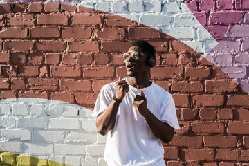 Young african american man dancing with headphones on colorful wall background. Concept of music, joy, freedom and urban lifestyle. Copy space